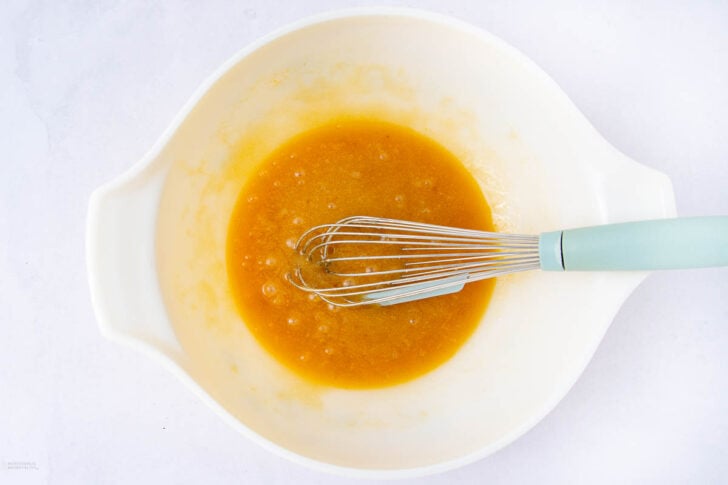 A white mixing bowl containing a yellow-orange liquid being whisked with a metal whisk that has a light blue handle, placed on a white surface.