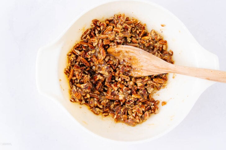 A white mixing bowl filled with a sticky pecan mixture, being stirred by a wooden spoon, on a white surface.