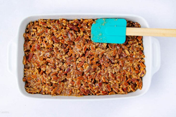A white rectangular baking dish filled with a pecan mixture, topped with chopped pecans, with a teal silicone spatula resting on the edge of the dish.