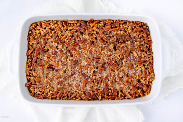 A white baking dish filled with a baked dessert topped with chopped pecans, set on a white cloth background. The topping appears golden brown and slightly crisp.