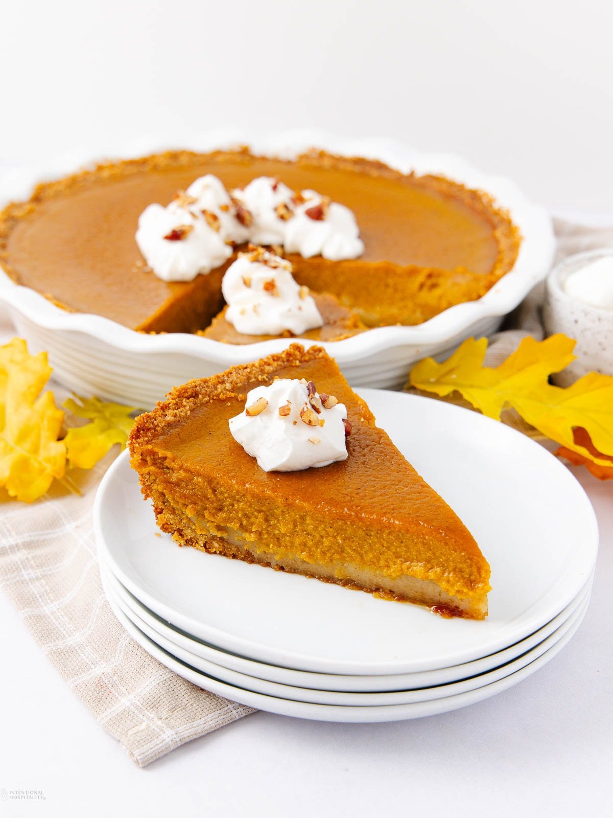A slice of pumpkin pie with a graham cracker crust and whipped cream sits on a stack of white plates. The rest of the pie, also topped with whipped cream and chopped nuts, is in the background. Yellow leaves surround the plates.