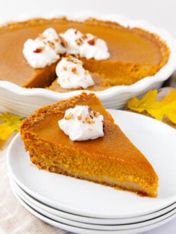 A slice of pumpkin pie topped with whipped cream and chopped nuts sits on a stack of white plates, with the rest of the pie and autumn leaves in the background.
