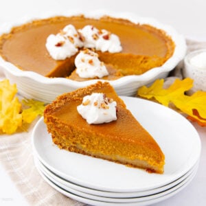 A slice of pumpkin pie topped with whipped cream and chopped nuts sits on a stack of white plates, with the rest of the pie and autumn leaves in the background.
