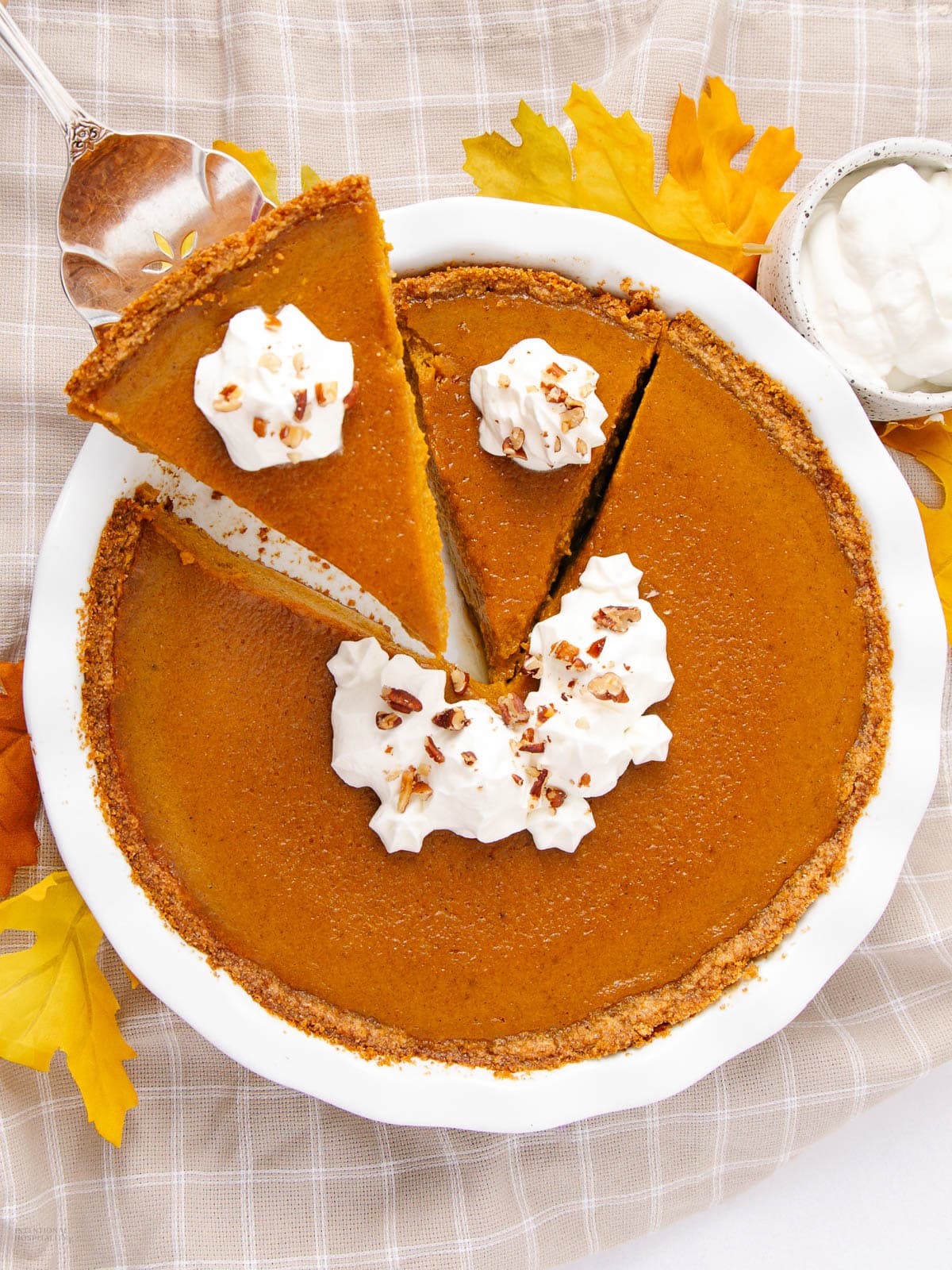 A pumpkin pie in a white dish with three slices cut, each topped with whipped cream and chopped nuts. A serving spoon holds one slice. Fall leaves and a bowl of whipped cream are nearby on a plaid cloth.
