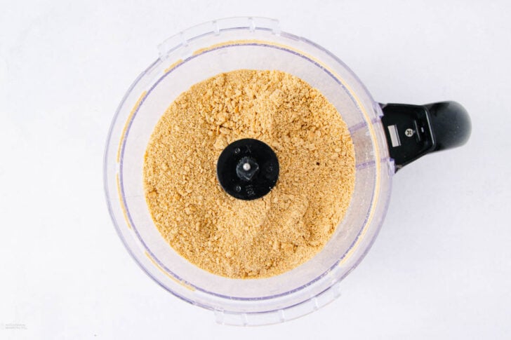 A food processor bowl filled with finely crushed golden-brown crumbs, viewed from above on a white background.