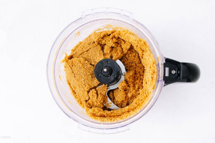 A food processor filled with finely ground, golden-brown crumbs, possibly graham cracker or cookie crumbs, forming a thick, even mixture around the blade. The appliance sits on a white surface.