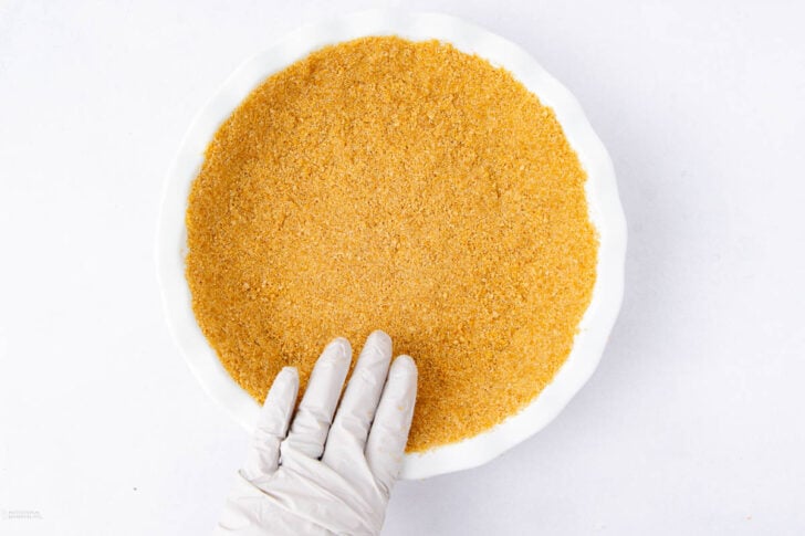 A gloved hand presses a layer of golden-brown graham cracker crumbs into a white pie dish on a white background.