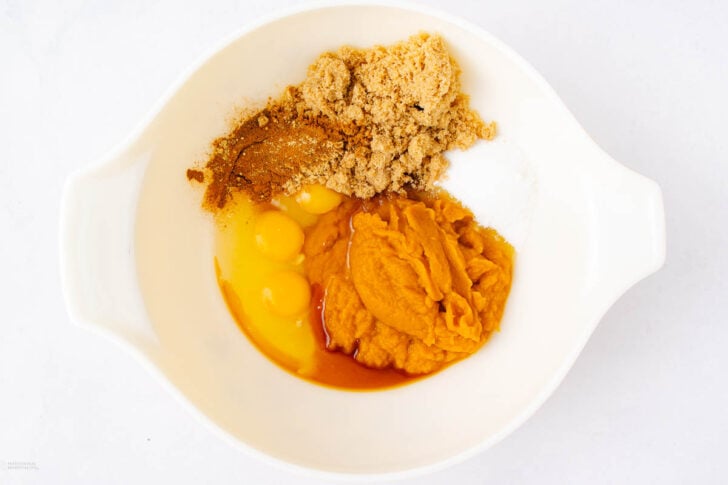 A white bowl containing eggs, brown sugar, pumpkin puree, ground spices, and baking powder or soda, seen from above on a white background.