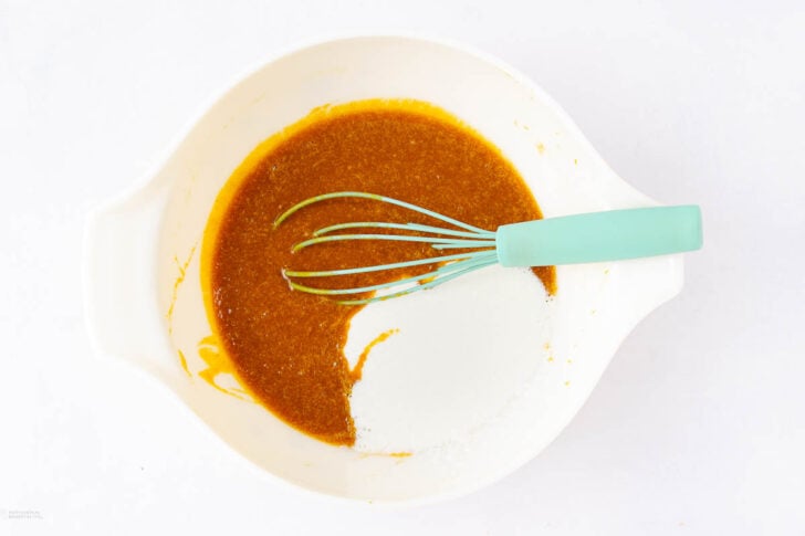 A white mixing bowl with brown liquid and white sugar inside, partially mixed with a teal-handled whisk, placed on a white surface.