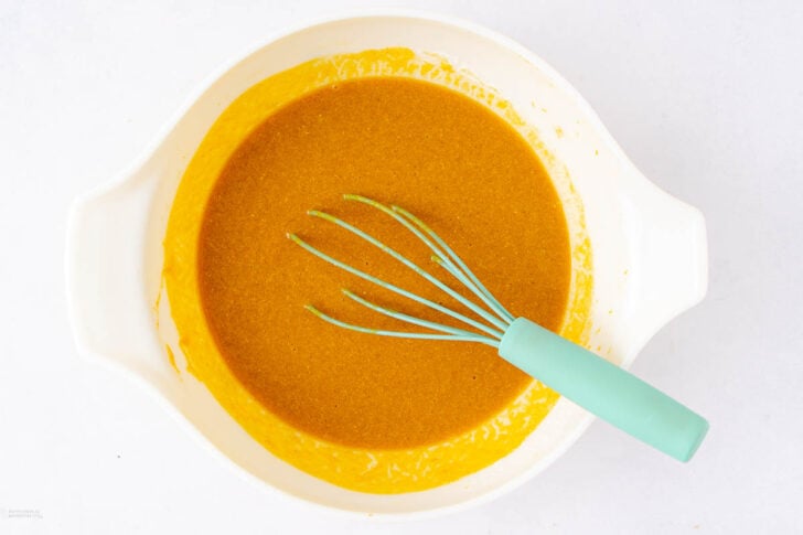 A white mixing bowl filled with a thick, golden-yellow batter. A light blue whisk with a green handle rests inside the bowl on the right side. The bowl is placed on a white surface.