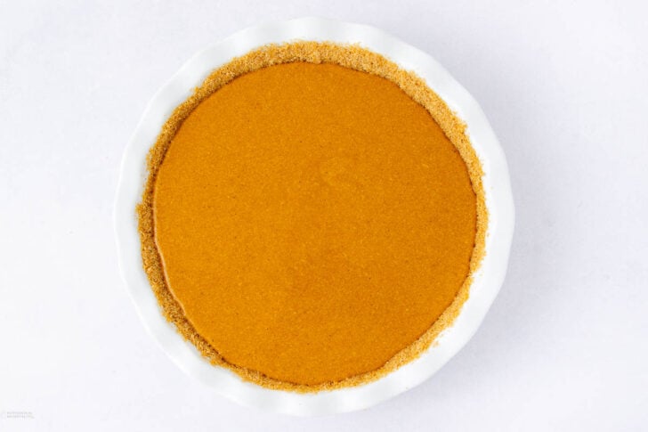 A round pie dish filled with a smooth, brownish-orange pumpkin pie filling, surrounded by a golden graham cracker crust, viewed from above on a white background.
