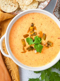 A bowl of creamy queso dip topped with chopped jalapeños and cilantro, placed next to tortilla chips, a wooden spoon, and a yellow napkin.