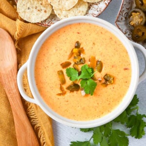 A bowl of creamy queso dip topped with chopped jalapeños and cilantro, placed next to tortilla chips, a wooden spoon, and a yellow napkin.