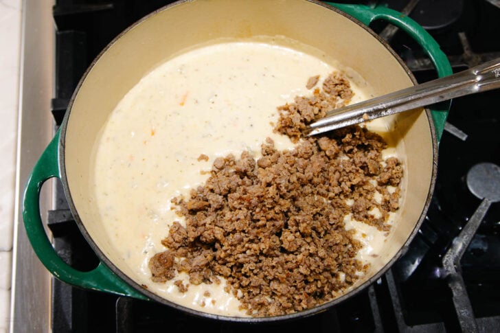 A green Dutch oven on a stovetop contains creamy soup being mixed with cooked ground meat using metal tongs.