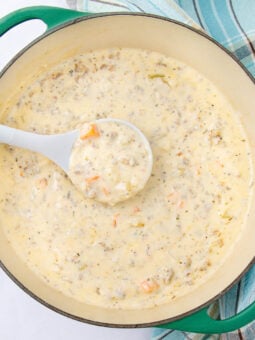 A creamy soup with chunks of vegetables and ground meat is being served from a green Dutch oven with a white ladle. A blue and white plaid kitchen towel is beside the pot.