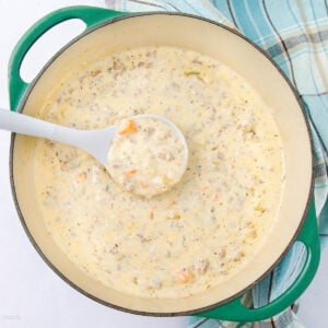 A creamy soup with chunks of vegetables and ground meat is being served from a green Dutch oven with a white ladle. A blue and white plaid kitchen towel is beside the pot.