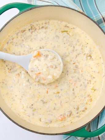 A creamy soup with chunks of vegetables and ground meat is being served from a green Dutch oven with a white ladle. A blue and white plaid kitchen towel is beside the pot.