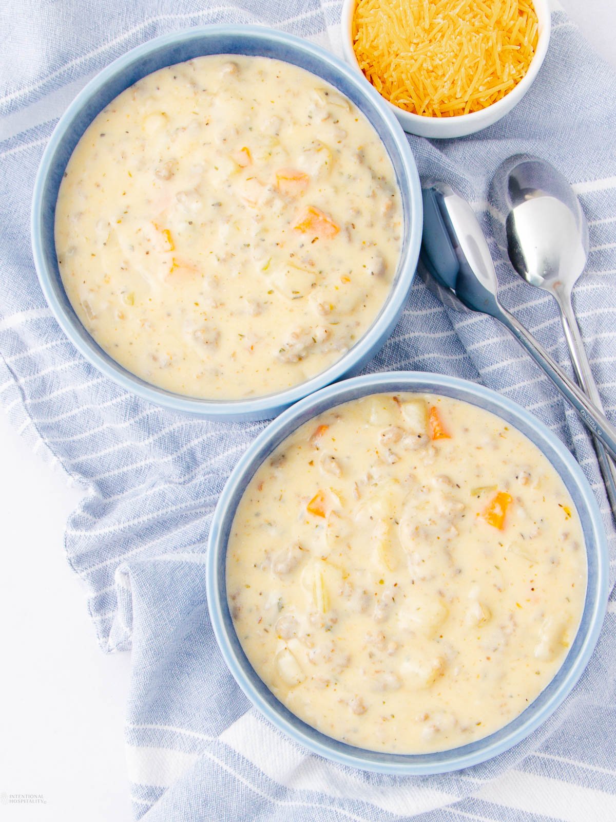 Two blue bowls filled with creamy soup containing visible chunks of sausage, potatoes, and vegetables sit on a blue striped cloth next to a small bowl of shredded cheese and three silver spoons.