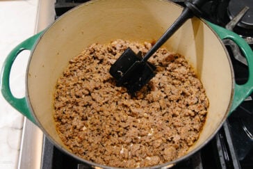 A green Dutch oven on a stove contains cooked and browned ground meat, being broken up with a black spatula.