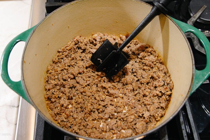 A green Dutch oven on a stove contains cooked and browned ground meat, being broken up with a black spatula.