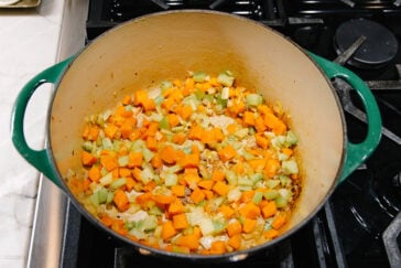 A large green Dutch oven on a stovetop contains diced carrots, celery, and onions being sautéed. The vegetables are partially cooked and starting to soften.