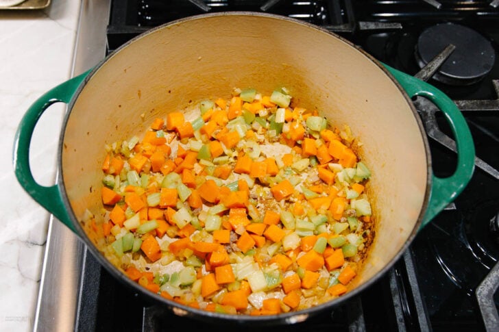 A large green Dutch oven on a stovetop contains diced carrots, celery, and onions being sautéed. The vegetables are partially cooked and starting to soften.