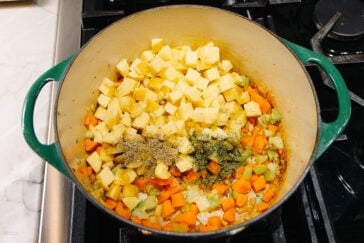 A green Dutch oven sits on a stove, filled with chopped potatoes, carrots, celery, onions, and herbs, ready to be cooked for a soup or stew.