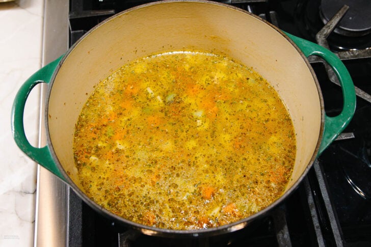 A green Dutch oven on a stove contains simmering vegetable soup with visible carrots, celery, and herbs in a golden broth.