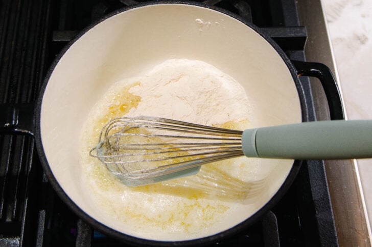 A whisk stirring flour and melted butter in a white pot on a stovetop, preparing a roux.