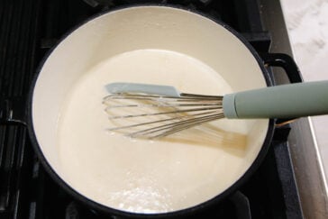A whisk with a green handle stirs a creamy white sauce in a large black pot on a stovetop.