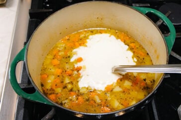 A green Dutch oven on a stovetop contains soup with diced vegetables and broth. A generous dollop of sour cream or cream is being stirred in with a metal spoon.