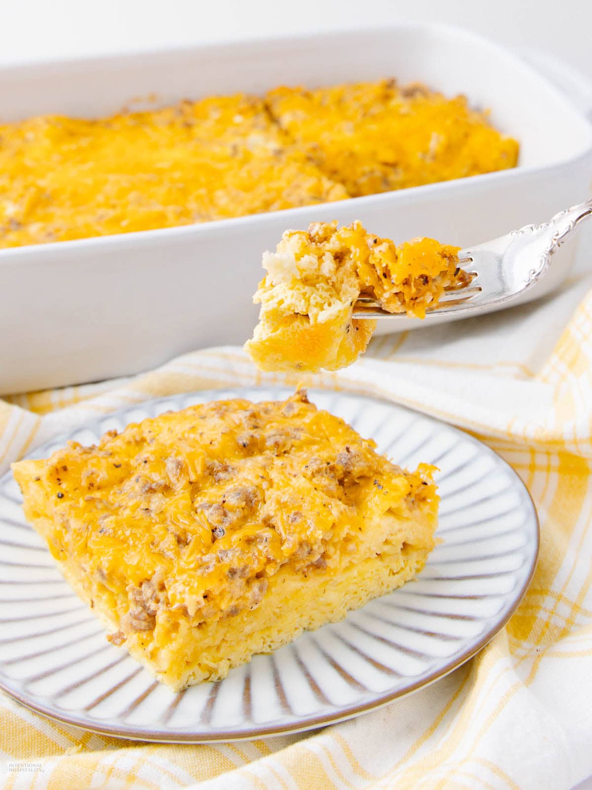 A square slice of cheesy breakfast casserole with ground sausage on a striped plate, with a fork holding a bite-sized piece above it. A baking dish with the remaining casserole is in the background.