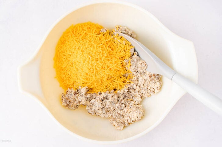 A white mixing bowl containing shredded cheddar cheese and a creamy mixture with ground beef, with a white mixing spoon resting inside.