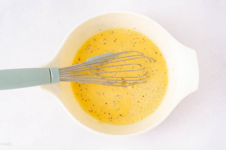 A whisk rests in a white mixing bowl filled with beaten eggs seasoned with black pepper, shown from above on a white surface.