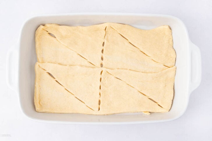A white baking dish with four pieces of uncooked crescent roll dough arranged to cover the bottom, with diagonal seams visible.