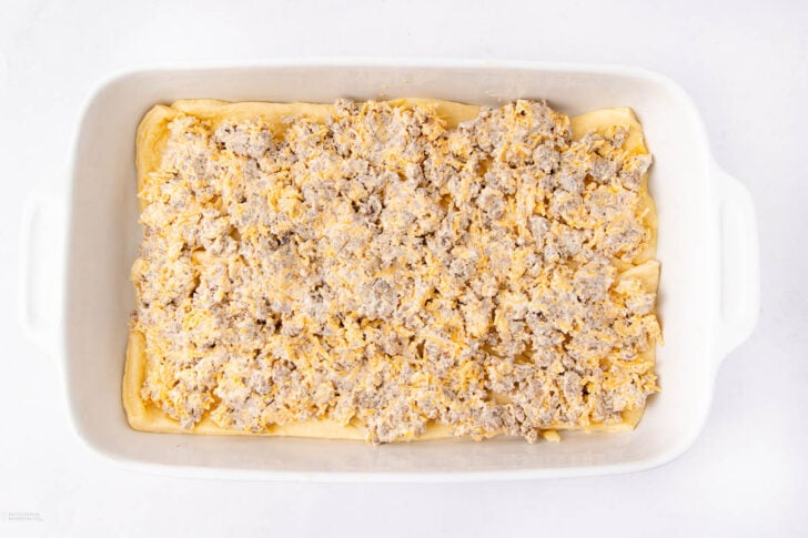 A white rectangular baking dish filled with an uncooked mixture of sausage, cheese, and dough spread evenly on the bottom. The dish is on a white surface.