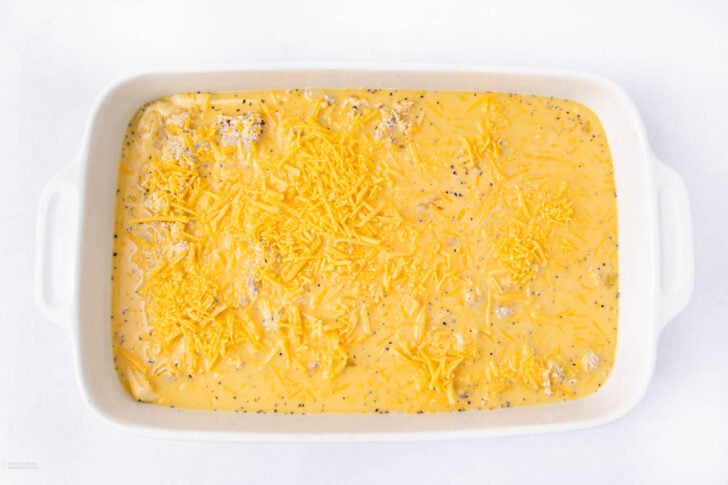 A white rectangular baking dish filled with an unbaked egg casserole mixture, topped with shredded cheddar cheese and visible ground sausage pieces, on a white surface.