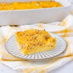 A slice of baked egg casserole with melted cheese and sausage is served on a striped plate. The rest of the casserole is visible in a white baking dish in the background, set on a yellow plaid cloth.