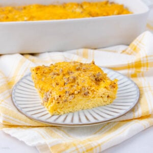 A slice of baked egg casserole with melted cheese and sausage is served on a striped plate. The rest of the casserole is visible in a white baking dish in the background, set on a yellow plaid cloth.