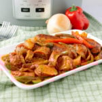 A rectangular white plate holds sliced sausages, onions, and bell peppers cooked in a red sauce. An onion, a red bell pepper, and a slow cooker are in the background on a green checkered cloth.