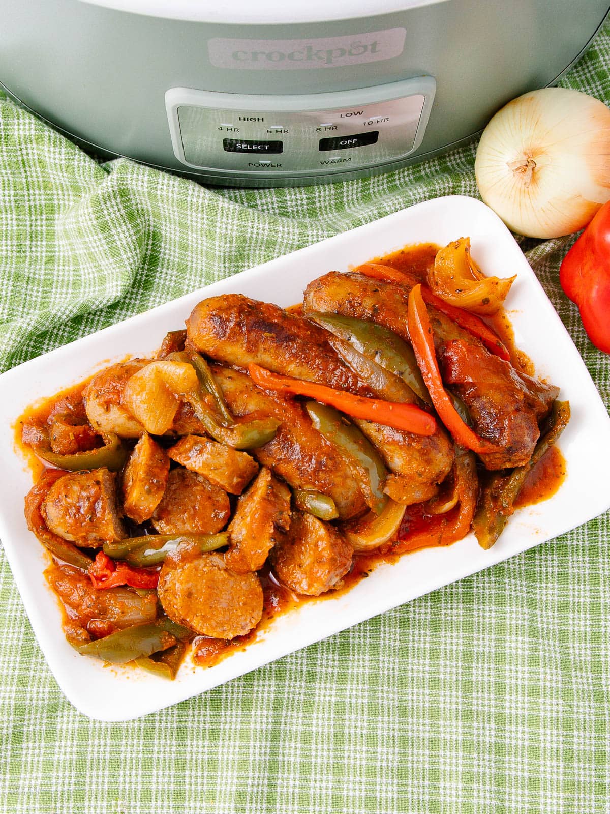 A white plate of cooked sausages with sliced bell peppers and onions in tomato sauce, placed on a green checkered cloth near a slow cooker, with a whole onion and bell pepper beside it.