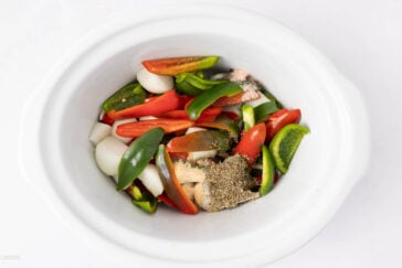 Sliced tomatoes, green bell peppers, onions, and seasonings are arranged in a white slow cooker, ready to be cooked.