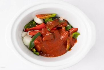 A white slow cooker filled with chopped green bell peppers, onions, tomatoes, and whole garlic cloves, all topped with a layer of red sauce.