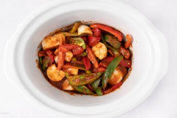 A white slow cooker filled with sliced green and red bell peppers, onions, and chunks of tomato in a red sauce, viewed from above on a white background.