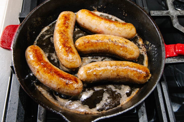 Five browned sausages cooking in a cast iron skillet with bubbling oil on a stovetop.