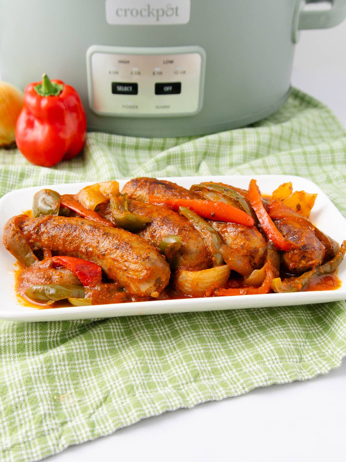 A white plate with cooked sausages, bell peppers, and onions in tomato sauce sits on a green checkered cloth, with a crockpot and fresh vegetables in the background.