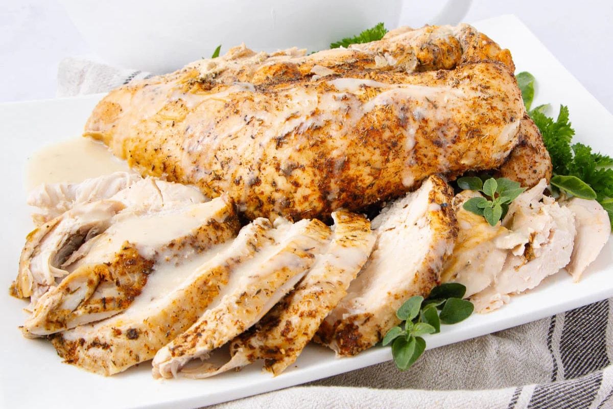 Sliced, seasoned turkey breast arranged on a white plate, garnished with fresh herbs and parsley. The turkey appears juicy and tender with visible spices and sauce on top.