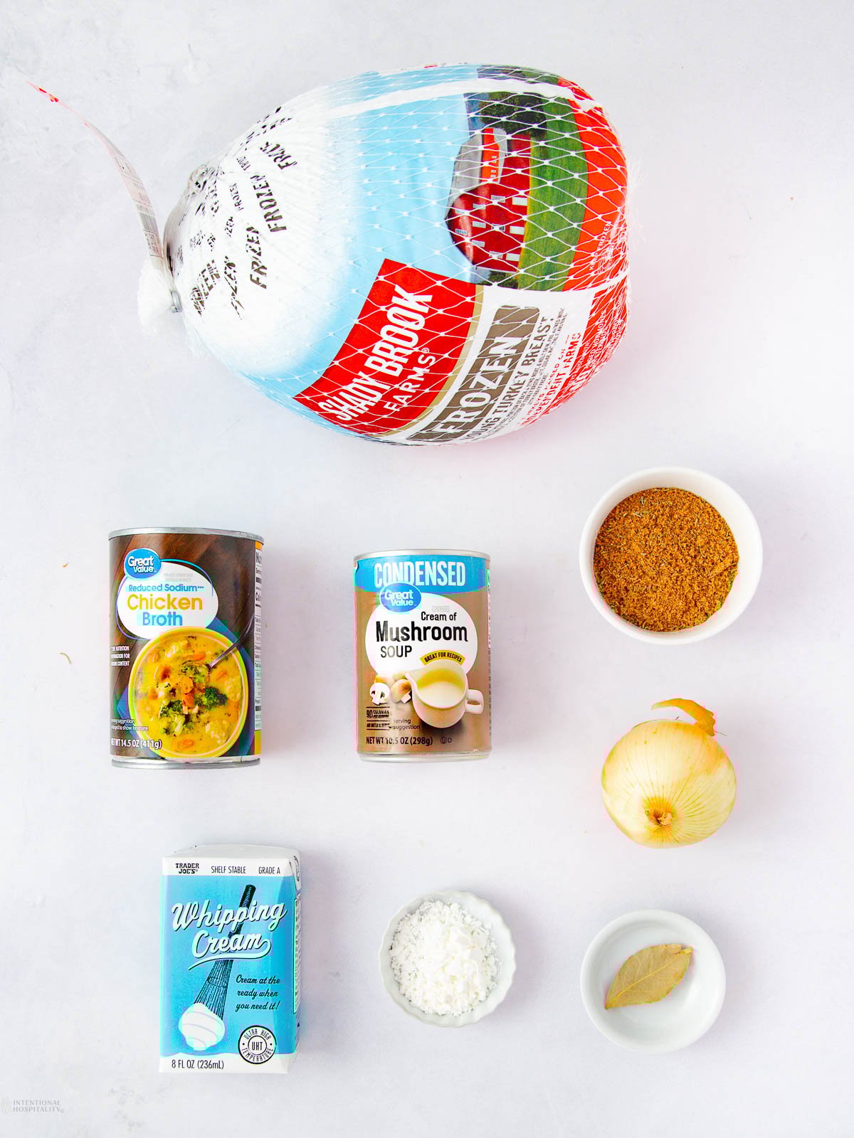 A frozen turkey, canned chicken broth, cream of mushroom soup, whipping cream, shredded coconut, brown seasoning, a yellow onion, and a bay leaf arranged on a white background.