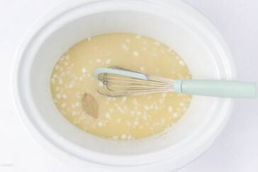 A white slow cooker contains a mixture of broth, chopped onions, a bay leaf, and a whisk with a light blue handle resting inside.