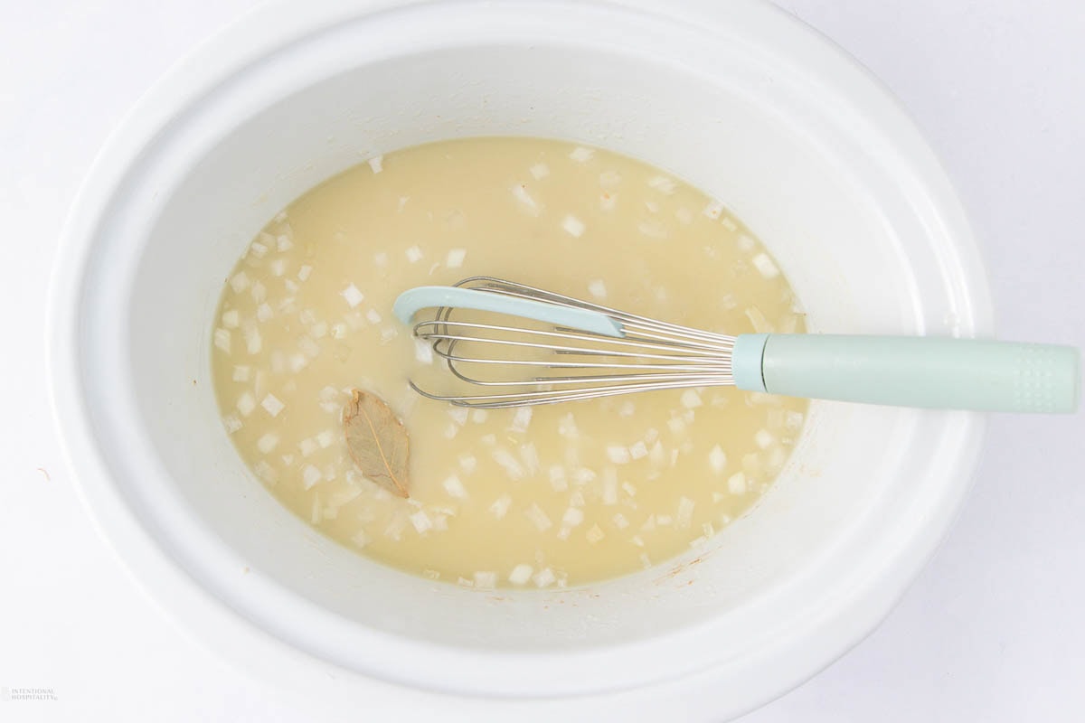 A white slow cooker contains a mixture of broth, chopped onions, a bay leaf, and a whisk with a light blue handle resting inside.